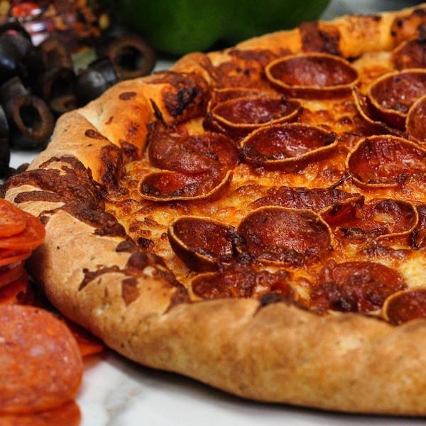 Close-up of a pepperoni pizza with a golden crust, alongside sliced pepperoni and black olives.