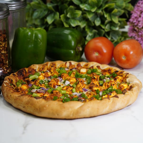 A chicken pizza with green onions and red onions, with bell peppers and tomatoes in the background.