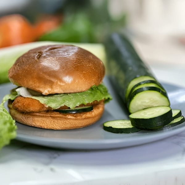 Chicken sandwich with lettuce and cucumber slices on a plate, with a whole cucumber in the background.