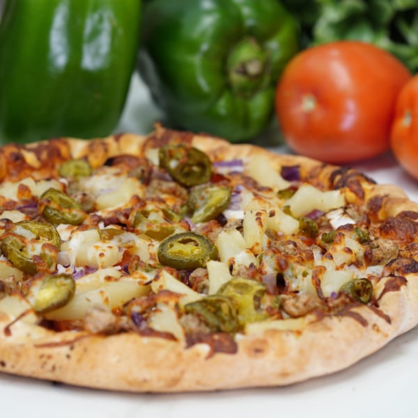 Slice Pizza & Krunchy Chicken pizza with pineapple, jalapeños, and sausage, with vegetables in the background.